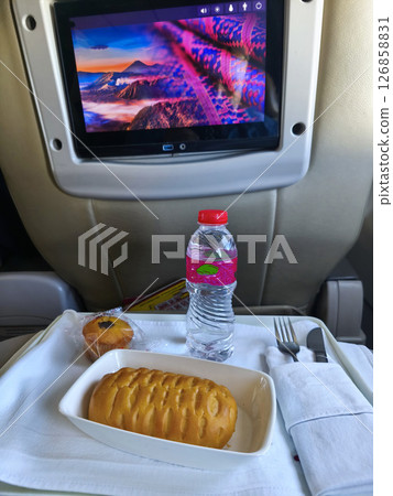 Airline meal with a water bottle and a muffin, viewed from a passenger's seat with an in-flight entertainment screen showing a colorful landscape. 126858831