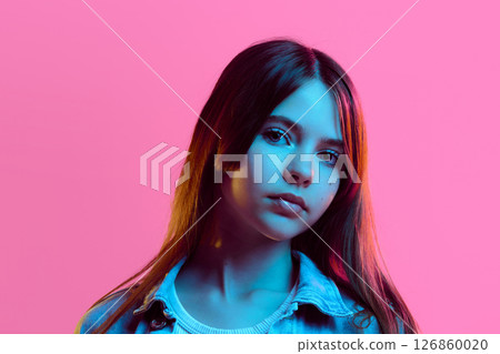Portrait of teen girl looking straight with calm expression under neon light, wearing denim jacket and white ribbed top, against pink gradient background 126860020