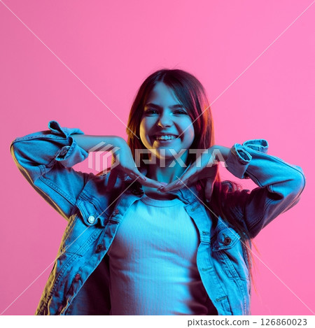 Teen girl smiling with hands raised under chin in playful gesture, wearing denim jacket and white top, against pink gradient background 126860023