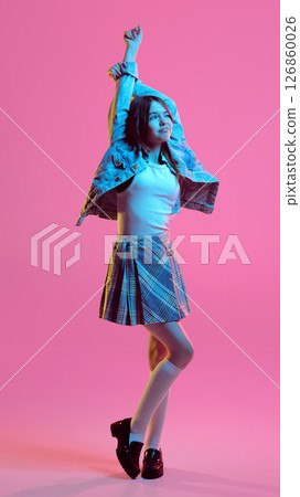 Teen girl stretching arms above head with soft smile in plaid skirt and denim jacket, against pink gradient background in neon light Teen girl stretching arms above head with soft smile in plaid skirt and denim jacket, against pink gradient background in neon light 126860026