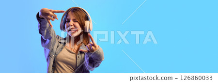 Banner. Teen girl making playful gestures and funny face in headphones while enjoying music against blue gradient background with negative space 126860033