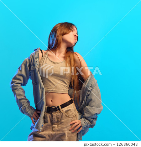 Teen girl with puckered lips and hands on hips poses confidently in crop top and denim under neon light against bright blue studio background. Teen girl with puckered lips and hands on hips poses confidently in crop top and denim under neon light against bright blue studio background. 126860048