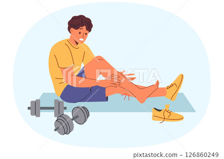 Injured sportsman sits on mat for sports near dumbbells, after stretching muscles on legs 126860249