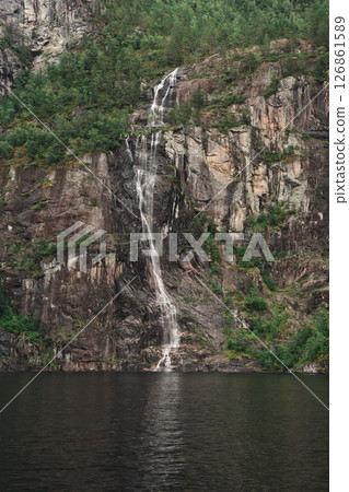 Thin fjord waterfall on dark cliff with forest near Bergen, Norway. 126861589