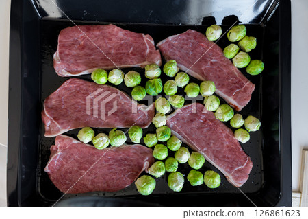 Raw pork steaks with Brussels sprouts arranged on black roasting tray for oven cooking 126861623