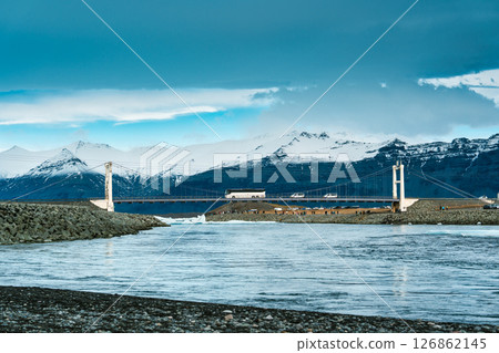 Iconic suspension bridge with car crossing over Jokulsarlon glacier lagoon and mountain range on background in Iceland Iconic suspension bridge with car crossing over Jokulsarlon glacier lagoon and mountain range on background in Iceland 126862145