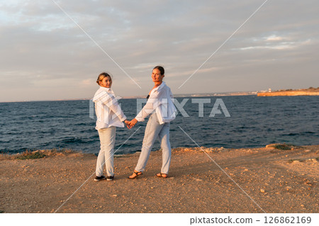 Couple Holding Hands Beach Sunset - Two people stand hand-in-hand on a beach at sunset. Couple Holding Hands Beach Sunset - Two people stand hand-in-hand on a beach at sunset. 126862169