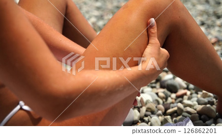 Woman beach cellulite. Woman checks cellulite on thigh, sitting at daytime on pebbled beach for body image. 126862201