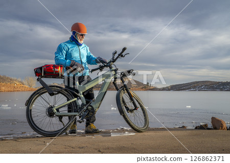 Senior cyclist with a touring e-bike with action camera mounted on handlebar at shore of frozen Horsetooth Reservoir in Colorado foothills. 126862371