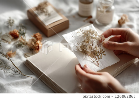 Close-up of hands arranging delicate dried flowers on paper, with an open journal and craft supplies in soft natural light. 126862865