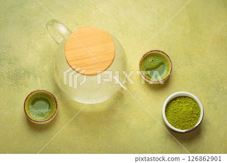 Steaming glass teapot and matcha green tea powder with two cups of prepared tea on green background top view Steaming glass teapot and matcha green tea powder with two cups of prepared tea on green background top view 126862901