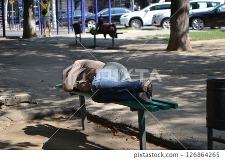 A homeless man is sleeping on a park bench or relaxing on a bench in an urban environment. Background with copy space for text. High quality photo 126864265