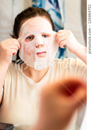 Young woman applying fabric face mask in bathroom mirror. Image for skincare routine, self care, and beauty treatment concepts. International Self Care Day. 126864701