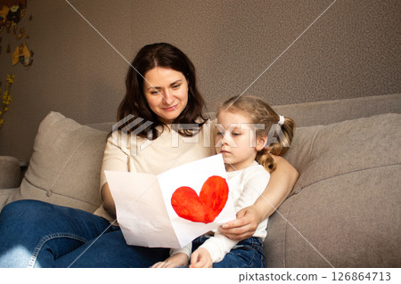 Little daughter with golden hair presents hand-drawn card to her smiling mother. Genuine moment of family bonding and innocent affection. Little daughter with golden hair presents hand-drawn card to her smiling mother. Genuine moment of family bonding and innocent affection. 126864713
