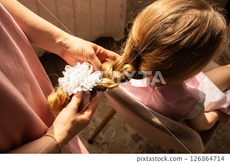 Blonde little girl sits patiently as mom carefully styles her hair into pretty braid for event. Heartwarming maternal care and childhood innocence. 126864714