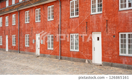 Colorful red buildings of Kastellet Barracks stand in Copenhagen, Denmark. This historic site features cobblestone pathways and charming architecture, inviting exploration of its rich history. 126865161