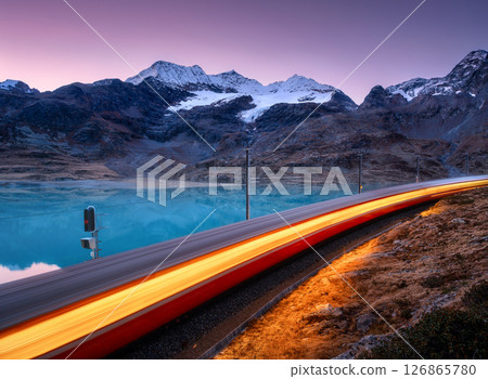 Moving red train near azure mountain lake, snowy rocks at sunset 126865780