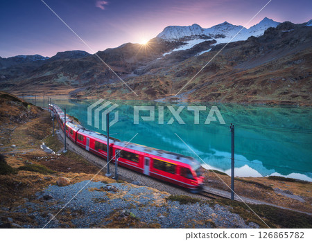 Moving red train near azure mountain lake, snowy rocks at sunset 126865782