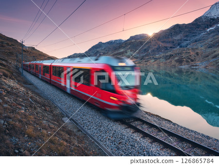 Moving red train near azure mountain lake, snowy rocks at sunset Moving red train near azure mountain lake, snowy rocks at sunset 126865784
