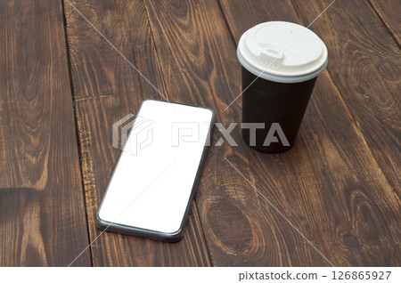 A smartphone with an empty screen and a paper coffee cup placed on a wooden table, offering a clean workspace. A smartphone with an empty screen and a paper coffee cup placed on a wooden table, offering a clean workspace. 126865927