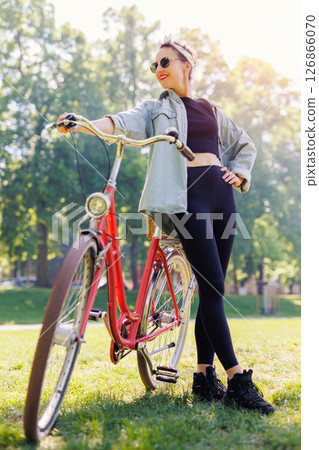 Portrait young adult pretty woman with red vintage retro bike and pin-up casual outfit green city park bright sunny summer day. Happy beautiful female person cycling outdoors. Healthy lifestyle 126866070