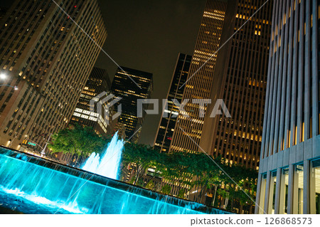At night in Manhattan, a vibrant blue fountain lights up a busy plaza surrounded by tall skyscrapers. People stroll by, enjoying the lively atmosphere of the urban environment. At night in Manhattan, a vibrant blue fountain lights up a busy plaza surrounded by tall skyscrapers. People stroll by, enjoying the lively atmosphere of the urban environment. 126868573