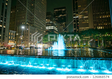 In Manhattan, a stunning fountain glows with blue light at night, surrounded by skyscrapers. People stroll along the water, enjoying the vibrant atmosphere of the city. In Manhattan, a stunning fountain glows with blue light at night, surrounded by skyscrapers. People stroll along the water, enjoying the vibrant atmosphere of the city. 126868574