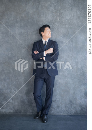 灰色背景中，一名身穿西裝、雙臂交叉的男子的全身 126870505