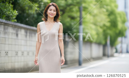 A woman in her 40s wearing a beige dress walking down a street full of greenery 126870525