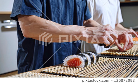 Professional chefs preparing salmon sushi rolls on bamboo mat, displaying precise culinary skills in contemporary kitchen workspace 126872098