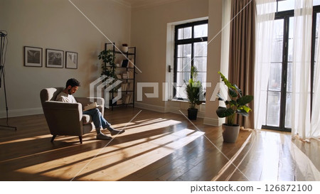 Young man sitting in armchair, reading book near large windows, surrounded by indoor plants in minimalist living room, experiencing peaceful afternoon 126872100