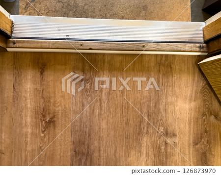 Close-up view of an interior door threshold showing the transition between wood flooring and a concrete surface. The metal strip and surrounding frame highlight the textures and construction details Close-up view of an interior door threshold showing the transition between wood flooring and a concrete surface. The metal strip and surrounding frame highlight the textures and construction details 126873970