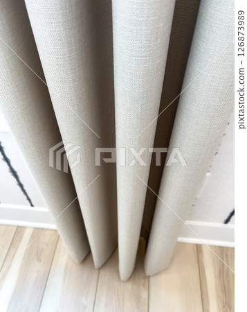 Vertical close-up of beige linen curtains hanging in soft pleats over a light wooden floor. The textured fabric and gentle lighting highlight the elegance and simplicity of modern interior design 126873989