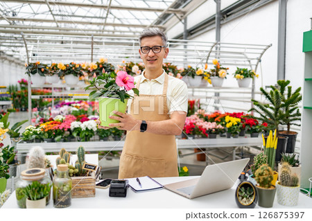 A Caucasian male florist stands in a shop holding a potted plant for sale. 126875397