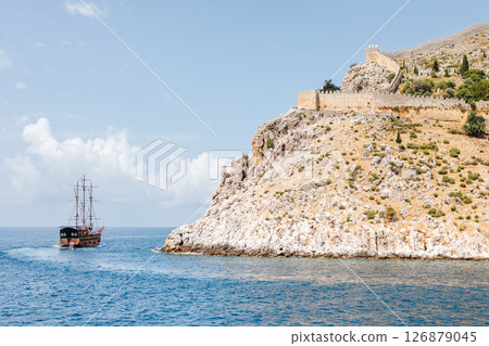 Old sailboat at the foot of a rocky coast with an ancient fortress wall on top. Beautiful seascape with a ship. Sea voyage Old sailboat at the foot of a rocky coast with an ancient fortress wall on top. Beautiful seascape with a ship. Sea voyage 126879045