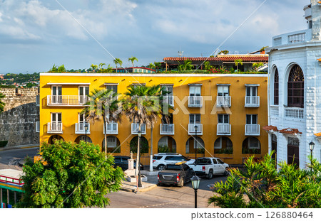 Vibrant street scene in Cartagena, Colombia featuring historic colonial buildings with arched windows, colorful facades, palm trees, and parked cars under a partly cloudy sky. Vibrant street scene in Cartagena, Colombia featuring historic colonial buildings with arched windows, colorful facades, palm trees, and parked cars under a partly cloudy sky. 126880464