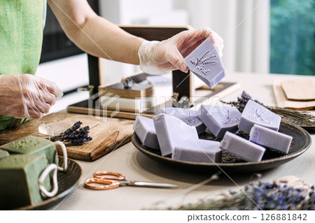 Person holds stamped lavender soap over a tray of handmade bars near a rustic soap station. Rustic wellness, handmade soap, small batch skincare, botanical fragrance Person holds stamped lavender soap over a tray of handmade bars near a rustic soap station. Rustic wellness, handmade soap, small batch skincare, botanical fragrance 126881842