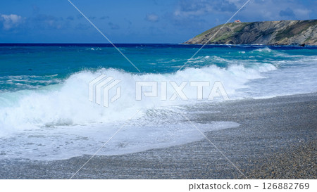 Powerful ocean waves crash onto a pebbled beach under a bright blue sky, with a rugged cliff and a distant lighthouse on a hill in the background 126882769