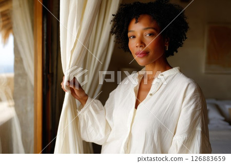 African American woman standing by window, holding curtain, natural light illuminating her face 126883659