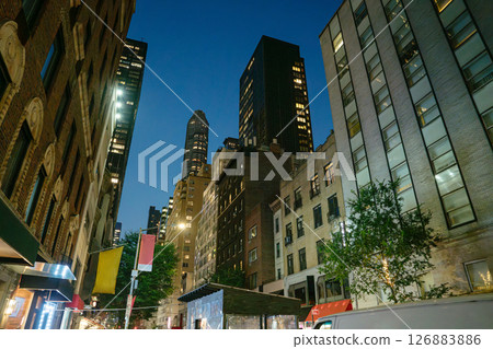 A quiet city street in Midtown Manhattan glows under warm lights as skyscrapers tower in the background. Residential buildings, banners, and a food kiosk line the street, creating a cozy urban A quiet city street in Midtown Manhattan glows under warm lights as skyscrapers tower in the background. Residential buildings, banners, and a food kiosk line the street, creating a cozy urban 126883886