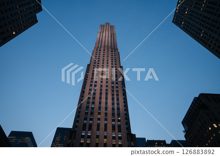 A dramatic upward view of Rockefeller Center captured at blue hour, framed by nearby skyscrapers. The fading daylight highlights the Art Deco architecture against a deep blue sky. 126883892