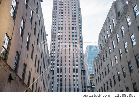 A centered view of the Rockefeller Center tower, framed tightly by symmetrical stone office buildings. The image highlights the Art Deco design and urban density of Midtown Manhattan. A centered view of the Rockefeller Center tower, framed tightly by symmetrical stone office buildings. The image highlights the Art Deco design and urban density of Midtown Manhattan. 126883893