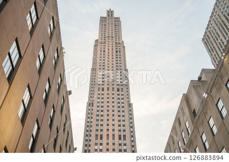 A striking view of the Rockefeller Center tower rising high between adjacent Art Deco buildings. The soft glow of the sunset illuminates the stone facade, highlighting the vertical symmetry and iconic 126883894