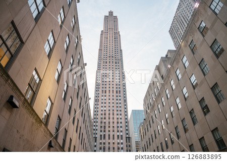 The towering Rockefeller Center rises between symmetrical Art Deco buildings in Midtown Manhattan. The iconic skyscraper stands tall under a soft, evening sky, framed by historic stone facades. The towering Rockefeller Center rises between symmetrical Art Deco buildings in Midtown Manhattan. The iconic skyscraper stands tall under a soft, evening sky, framed by historic stone facades. 126883895