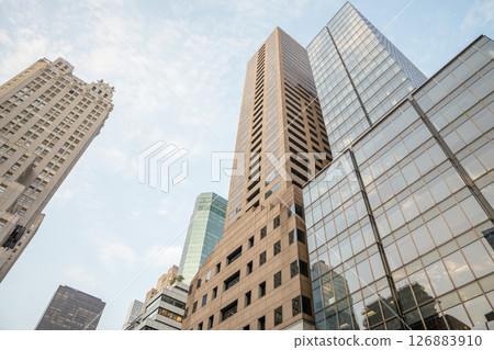 Modern high-rise office buildings reach into the sky in Midtown Manhattan. Reflective glass windows mirror the blue sky and clouds, creating a dynamic urban scene. Modern high-rise office buildings reach into the sky in Midtown Manhattan. Reflective glass windows mirror the blue sky and clouds, creating a dynamic urban scene. 126883910