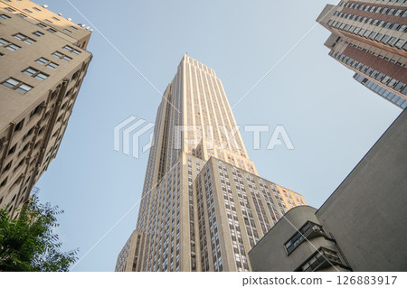 The Empire State Building towers above the surrounding city structures. Photographed from a low angle, the skyscraper appears majestic against the clear blue sky. 126883917