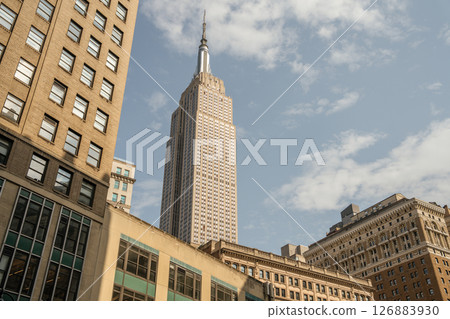 The iconic Empire State Building towers above surrounding structures. The photo captures its pointed spire and detailed windows on a clear day. 126883930
