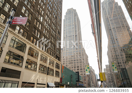 Tall buildings line a narrow street in Midtown Manhattan. One facade reflects the skyline in its mirrored glass. 126883935