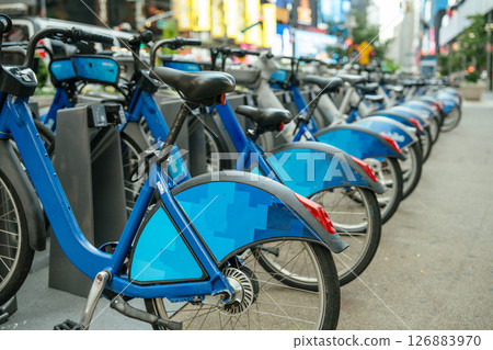 A row of blue public rental bikes is neatly lined up at a docking station on a busy city sidewalk. The background reveals blurred traffic, lights, and tall buildings, suggesting a vibrant urban A row of blue public rental bikes is neatly lined up at a docking station on a busy city sidewalk. The background reveals blurred traffic, lights, and tall buildings, suggesting a vibrant urban 126883970