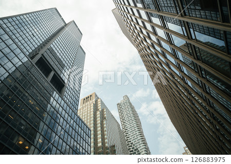 An upward view of modern high-rise office buildings with glass facades and reflective windows. The photo captures a dramatic city perspective under a bright, slightly cloudy sky, showcasing urban An upward view of modern high-rise office buildings with glass facades and reflective windows. The photo captures a dramatic city perspective under a bright, slightly cloudy sky, showcasing urban 126883975